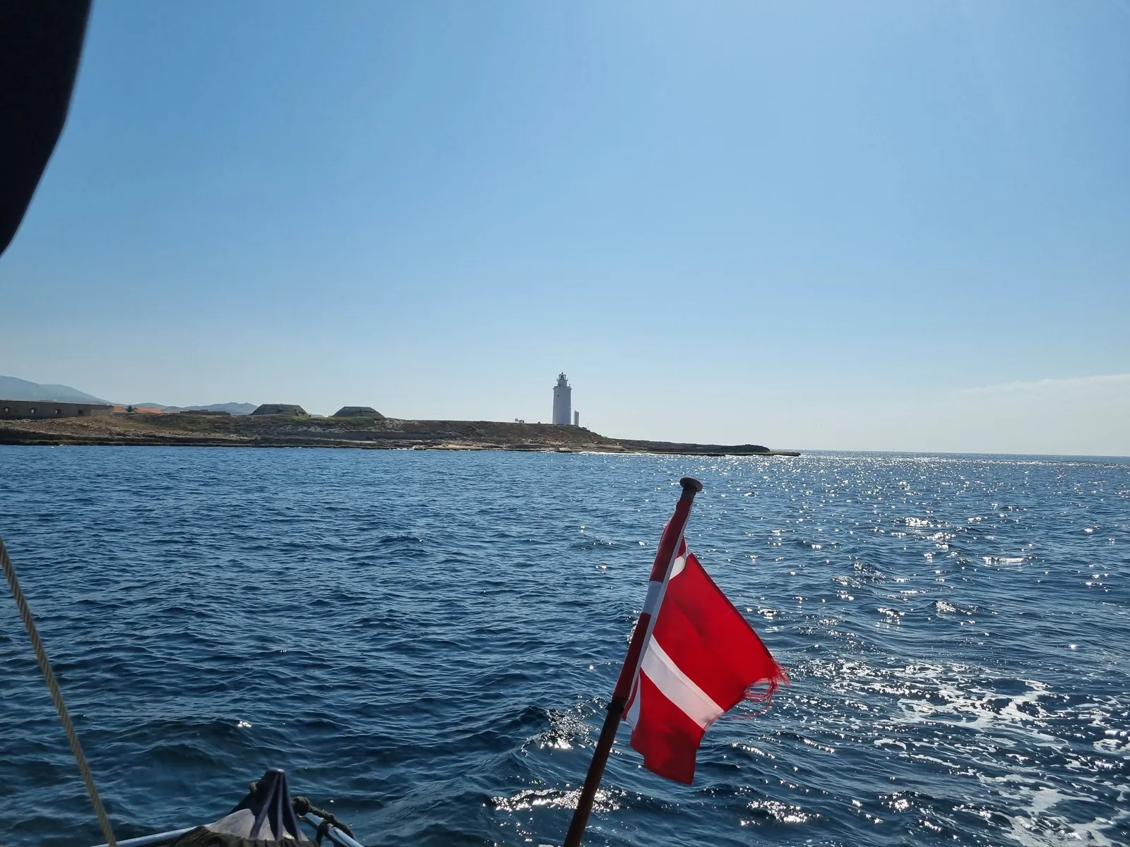 Danish flag on the stern of a Hanse 575 passing a lighthouse in the Strait of Gibraltar during a yacht delivery, June 2026 Danish flag on the stern of a Hanse 575 passing a lighthouse in the Strait of Gibraltar during a yacht delivery, June 2026